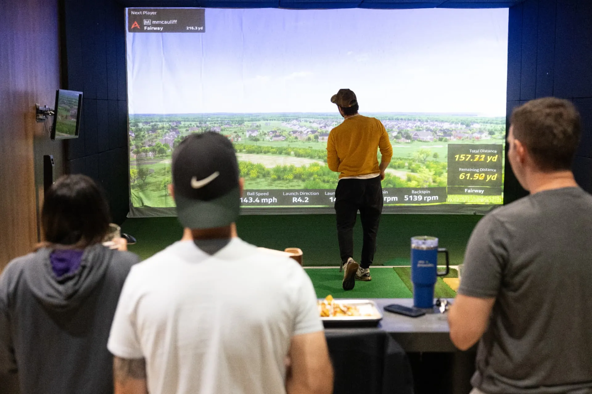 Friends watching a golf shot in a simulator bay with drinks and shared snacks on the counter