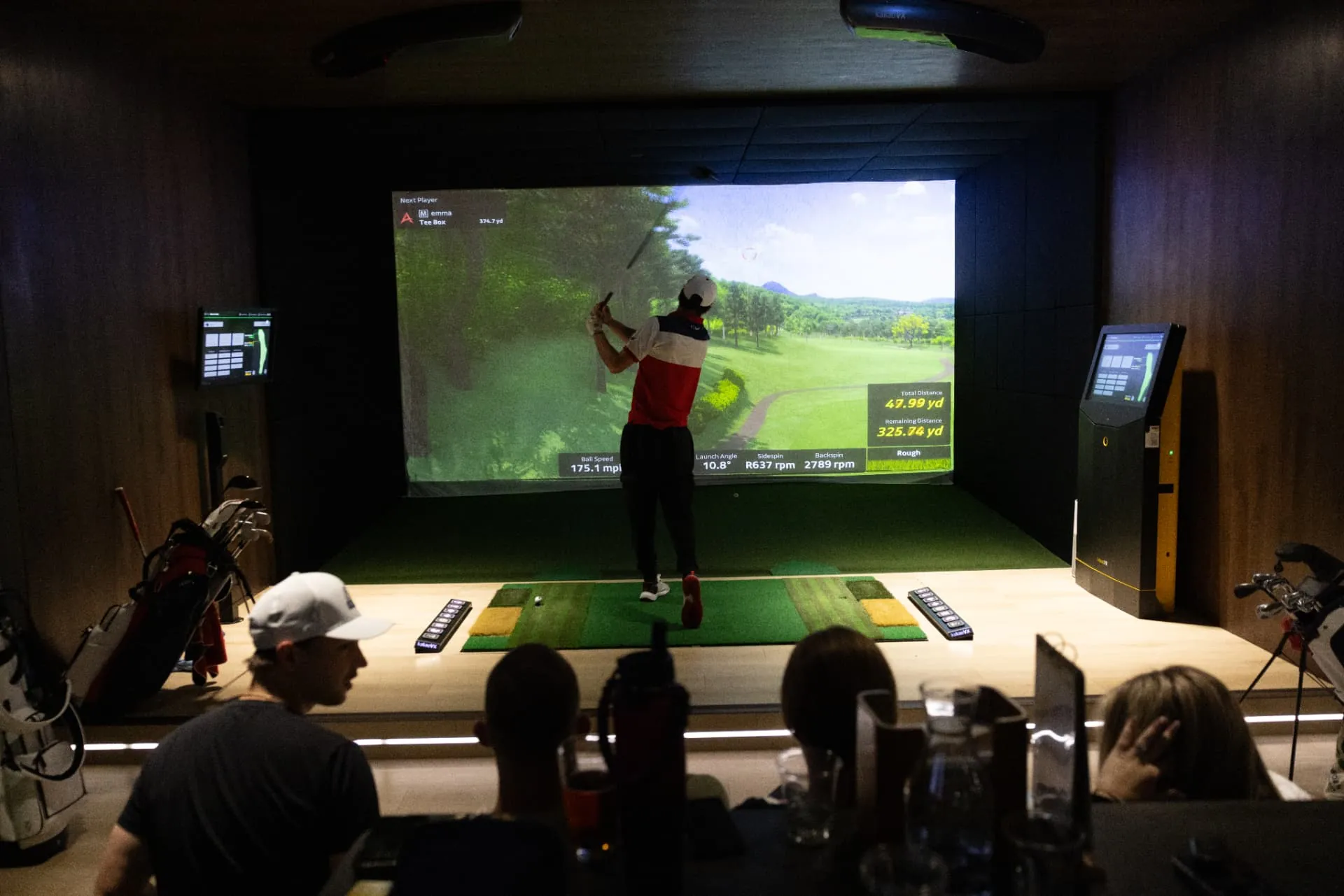 Golfer taking a swing in an indoor simulator bay while seated friends watch the shot on the course screen